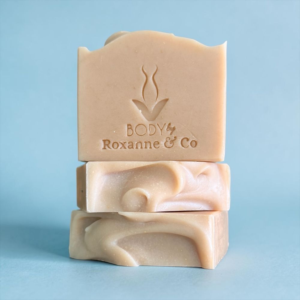 Three stacked handmade soap bars in a light peach-beige color, with the top bar embossed with the brand name 'BODY Roxanne & Co', set against a solid light blue background.