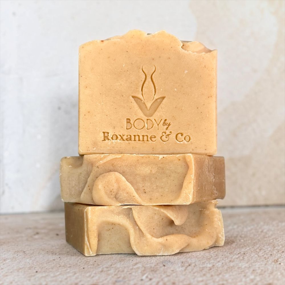 Three stacked handmade soap bars in a warm sandy beige color, with the top bar embossed with the brand name 'BODY by Roxanne & Co', set against a neutral textured background.