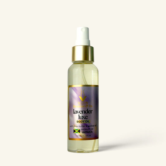 A bottle of Lavender Luxe Body Oil by Sweetener & Co. The clear bottle shows a pale yellow oil inside, with a white spray top, a metallic gold collar, and a purple label featuring gold and black text.
