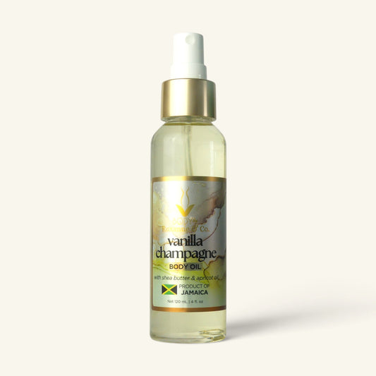 A clear glass bottle of Vanilla Champagne Body Oil with a white spray top and gold collar, featuring a gold and green label, set against a solid off-white background.