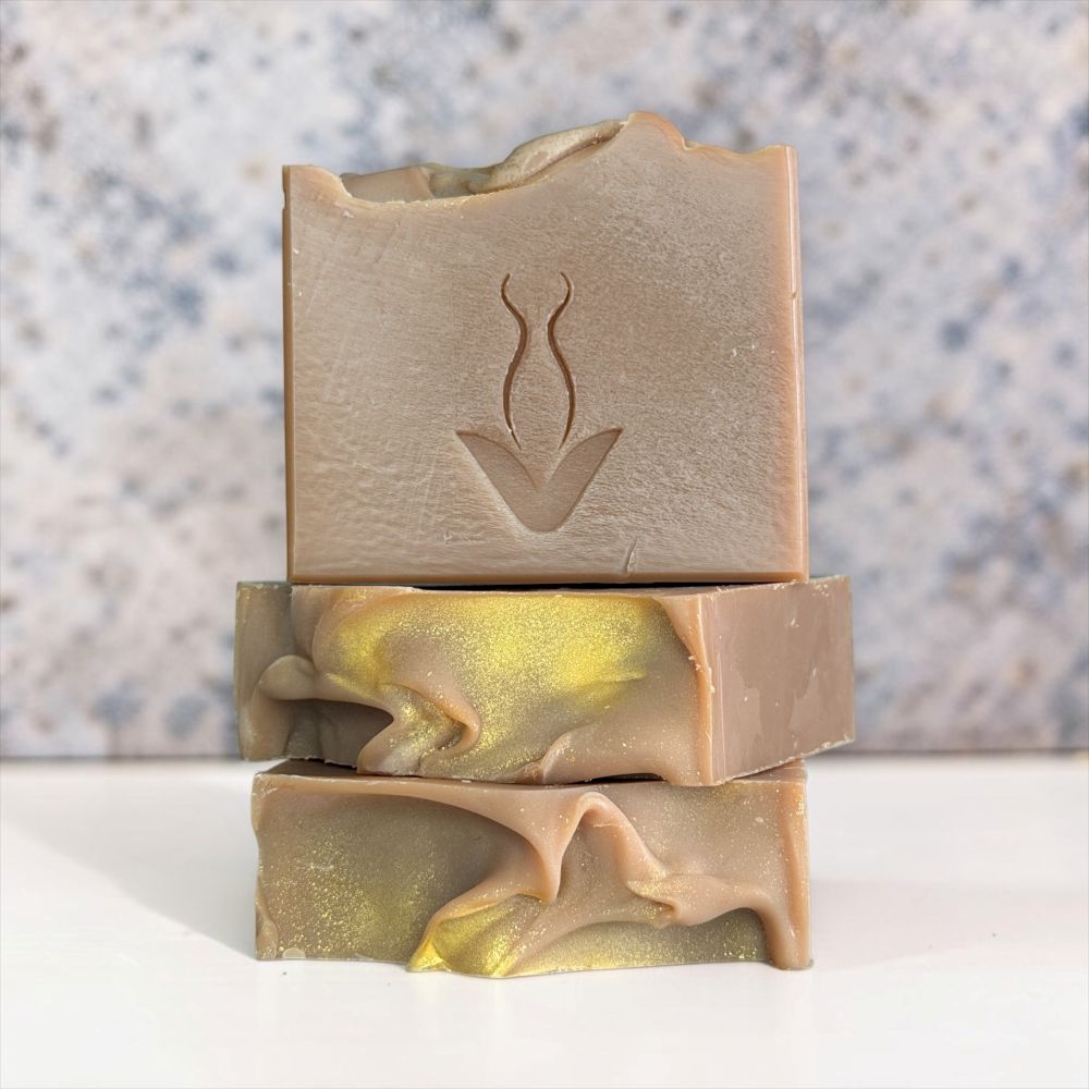Three stacked handmade soap bars with a swirled beige and gold marbled pattern, featuring an embossed botanical logo on the top bar, set against a speckled gray background.