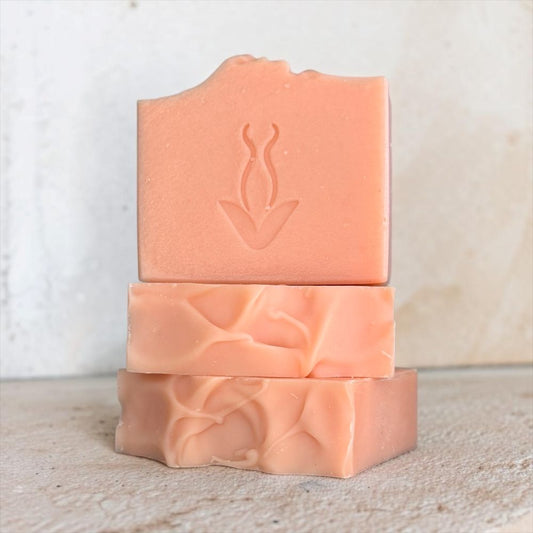 Three stacked rectangular bars of handmade soap in a soft peach-pink color, with a debossed botanical logo on the top bar, set against a neutral textured light background.