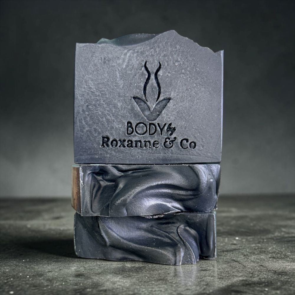 A stacked set of dark gray artisan soap bars with a textured top bar embossed with the brand 'BODY by Roxanne & Co' and a stylized plant logo, set against a dark gray background.