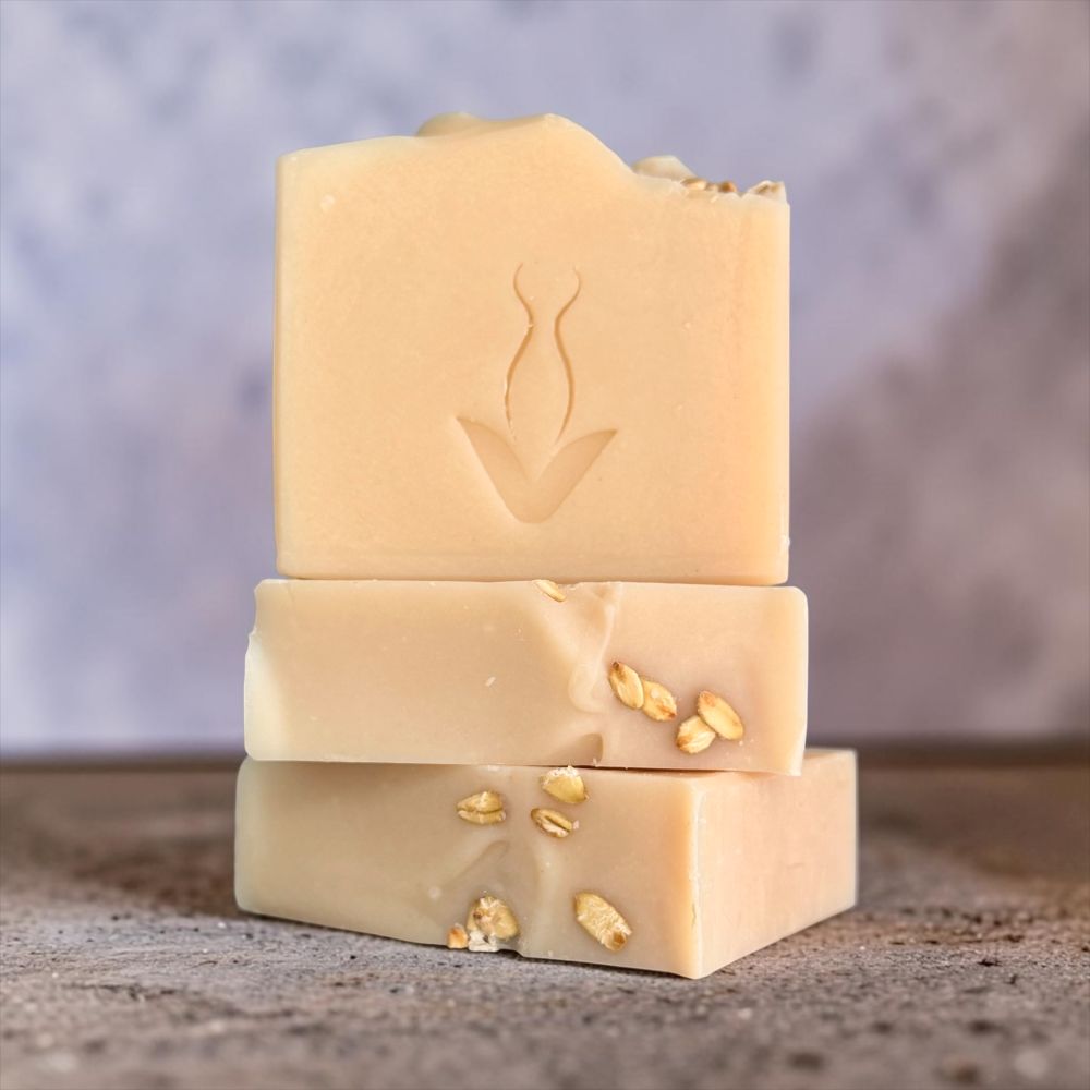Three stacked handmade soap bars in warm cream and light tan shades, each embedded with small dried botanical pieces, set against a soft textured gray background.