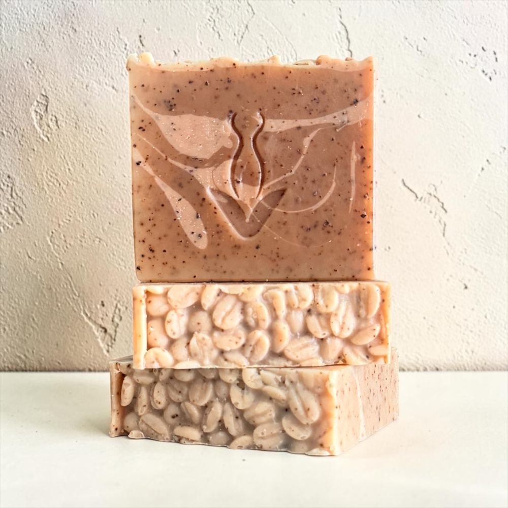 Three stacked handmade soap bars with speckled oatmeal tones and embedded botanical elements, set against a textured off-white background.