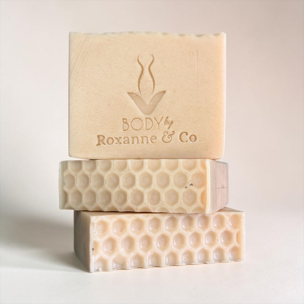 Three stacked handmade soap bars in a warm cream-beige color, with the top bar embossed with the brand name 'BODY by Roxanne & Co' and a simple logo. The soaps feature different textured patterns, including a honeycomb design on the bottom two bars, set against a clean, light beige background.