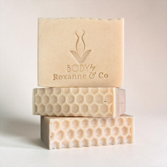 Three stacked handmade soap bars in a warm cream-beige color, with the top bar embossed with the brand name 'BODY by Roxanne & Co' and a simple logo. The soaps feature different textured patterns, including a honeycomb design on the bottom two bars, set against a clean, light beige background.