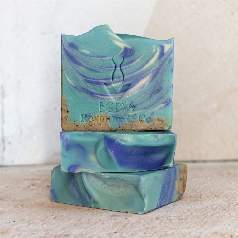Three stacked handmade soap bars with a swirled teal, blue, and purple pattern and a natural rough bottom edge. The top bar is embossed with the brand name 'BODY by Roxanne Co.