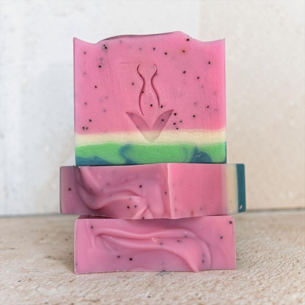 Three stacked handmade soap bars with a pink top layer, a green and blue marbled middle layer, and small black speckles, set against a neutral textured background.