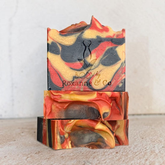 Three stacked handmade soap bars with a marbled swirl pattern of red, orange, yellow, and black, featuring the brand name 'BODY by Roxanne & Co' embossed on the top bar.