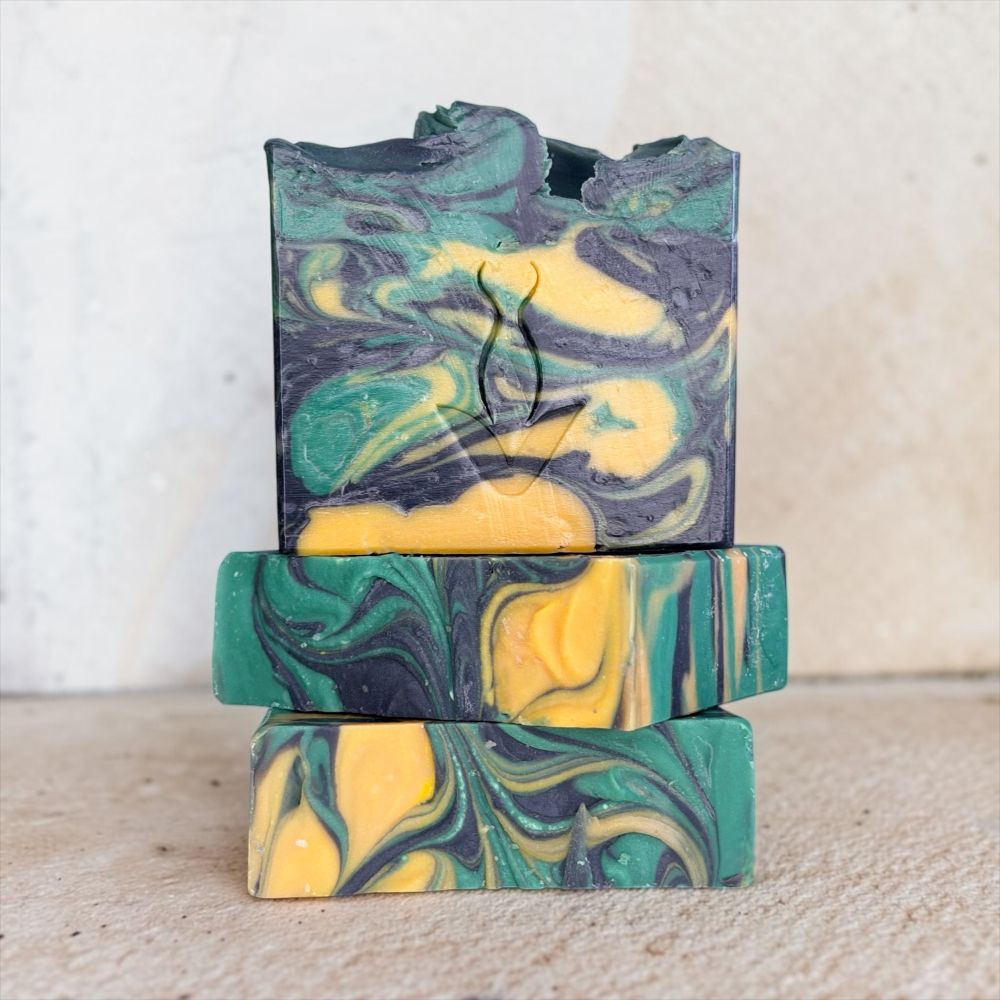 Three stacked handmade soap bars with a marbled swirl pattern of green, yellow, and dark gray, photographed against a neutral light beige background.