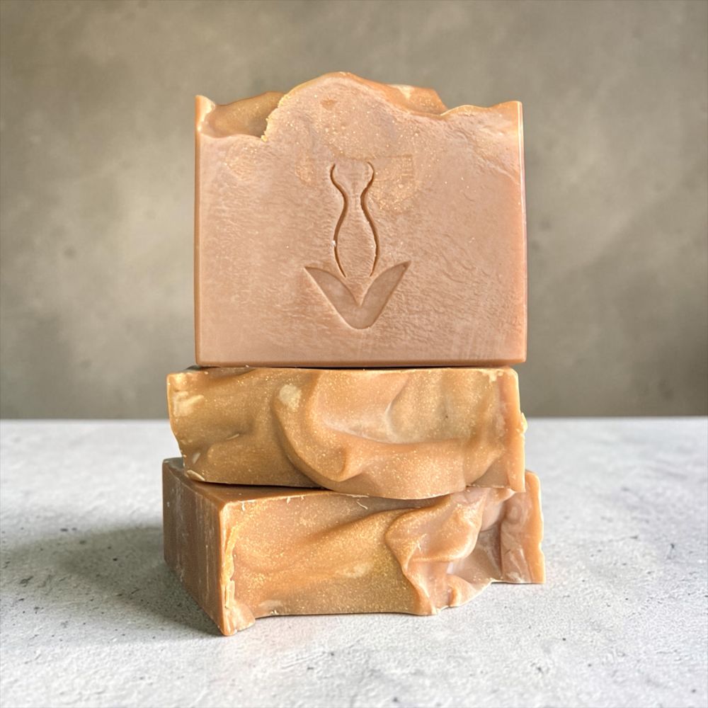Three stacked handmade soap bars in warm peach and tan shades, with the top bar embossed with a stylized tulip design, set against a neutral gray background.