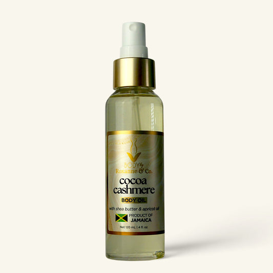 A clear spray bottle of Roxanne & Co Cocoa Cashmere Body Butter with a white pump and gold collar, containing a pale yellow liquid, set against a solid off-white background.
