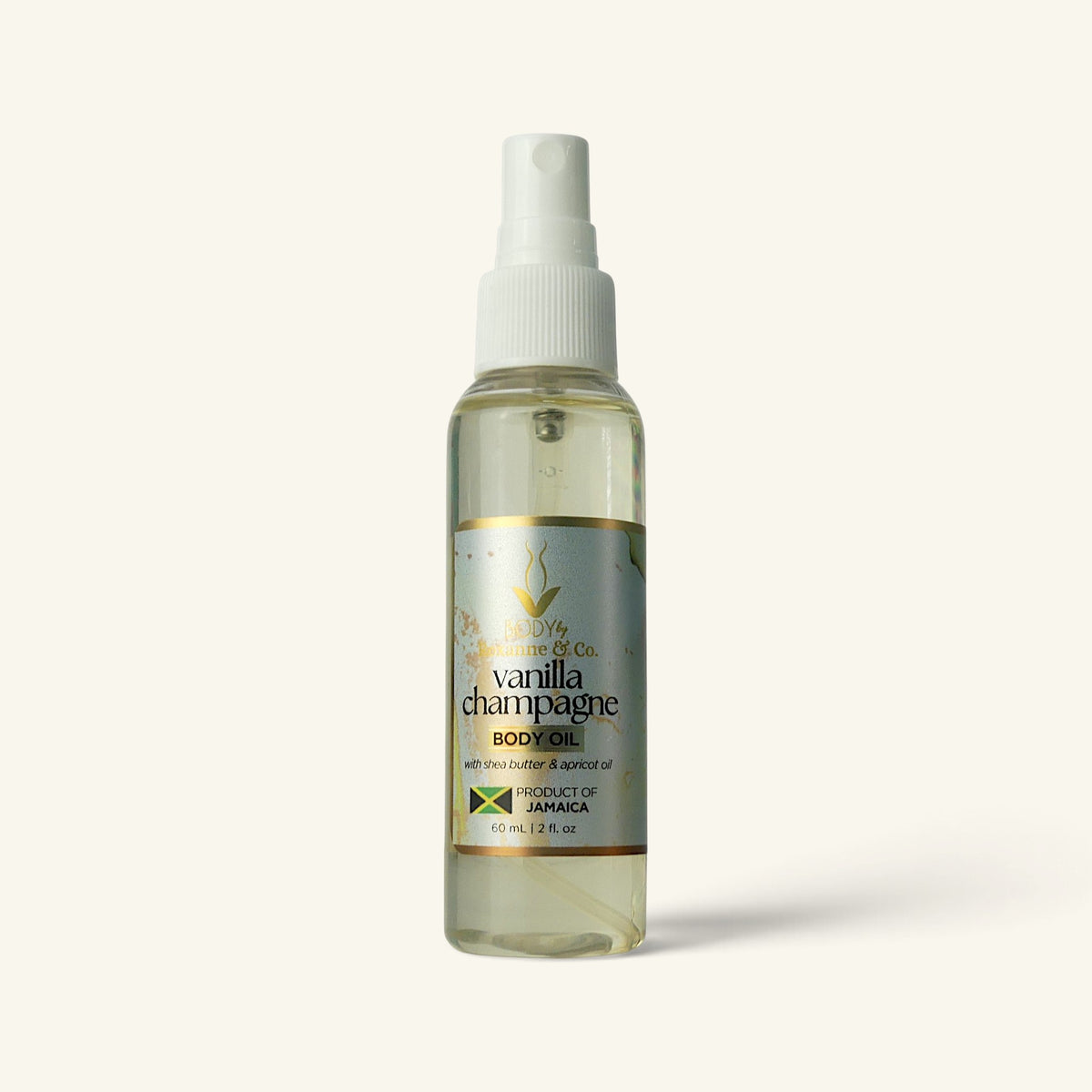 A bottle of Vanilla Champagne Body Oil by The Body of Co. The clear spray bottle shows a pale yellow oil inside, with a white pump dispenser and a silver and gold label.