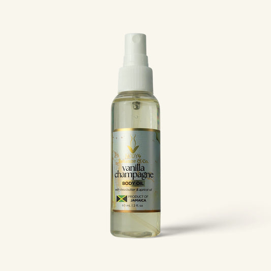 A bottle of Vanilla Champagne Body Oil by The Body of Co. The clear spray bottle shows a pale yellow oil inside, with a white pump dispenser and a silver and gold label.