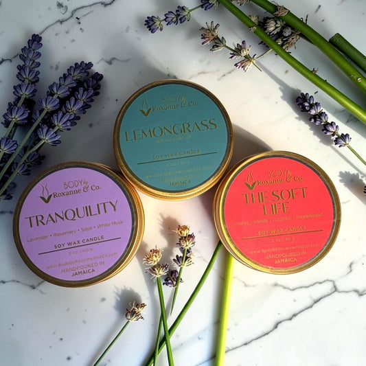 Three scented candle tins with labels 'Lemongrass', 'Tranquility', and 'The Soft Life' on a marble surface with lavender and bamboo.