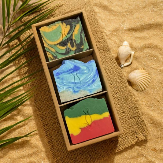 Three colorful soap bars in a box on a sandy surface with shells and palm leaves.