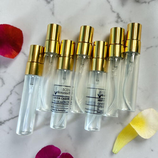 Six clear glass perfume atomizer travel spray bottles with gold caps, arranged on a white marble surface with scattered rose petals. Two bottles have visible white labels reading 'BOOCHEN Roxanne' and 'COCOA CASHMERE'.