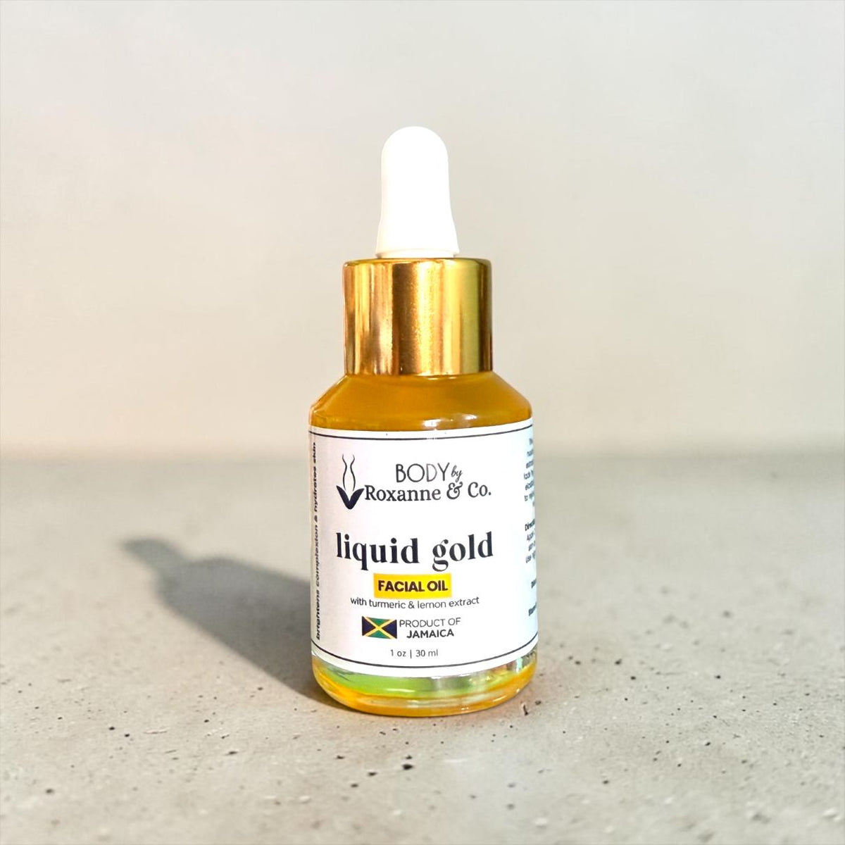 A small glass dropper bottle of BODY by Roxanne & Co. Liquid Gold Facial Oil, filled with golden-yellow oil and featuring a white label with a gold cap and white dropper top, set on a speckled gray surface against a light beige background.