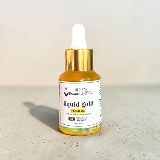 A small glass dropper bottle of BODY by Roxanne & Co. Liquid Gold Facial Oil, filled with golden-yellow oil and featuring a white label with a gold cap and white dropper top, set on a speckled gray surface against a light beige background.