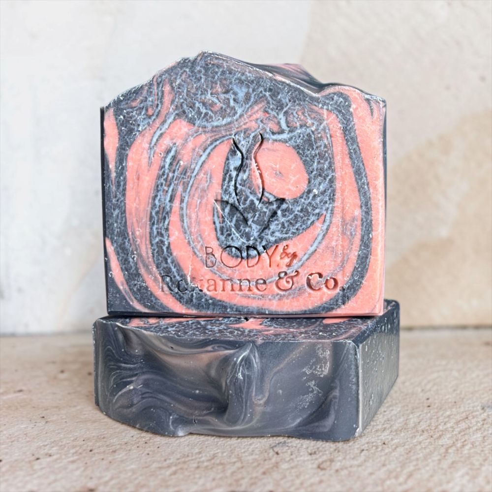 A handmade soap bar with a marbled swirl pattern of dark charcoal gray and dusty pink, stacked on a matching dark gray bar. The top bar features an embossed design of a flame and the text 'BODY &ianne & Co'.